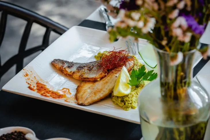  a white plate of sea bass fillet with vegetables, a vase of flowers next to it on a table 