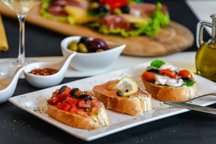  a white plate with three pieces of bruschetta on a table, olive oil on the side and platter of cheese and ham in the background